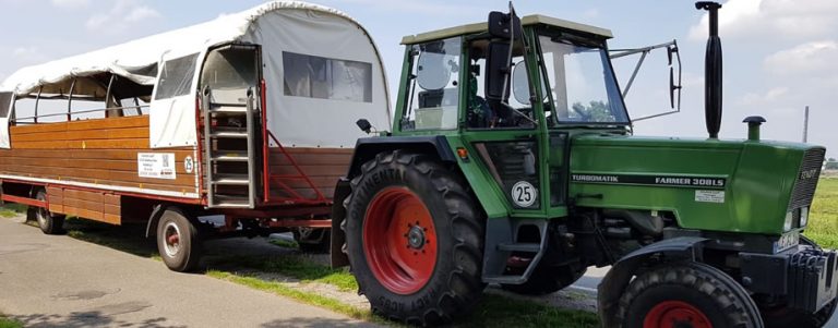 Unser Planwagen am Haken eines grünen Traktors: hölzerner Aufbau mit heller Plane, seitlichem Einstieg und Treppe, aufgenommen auf einem Feldweg.